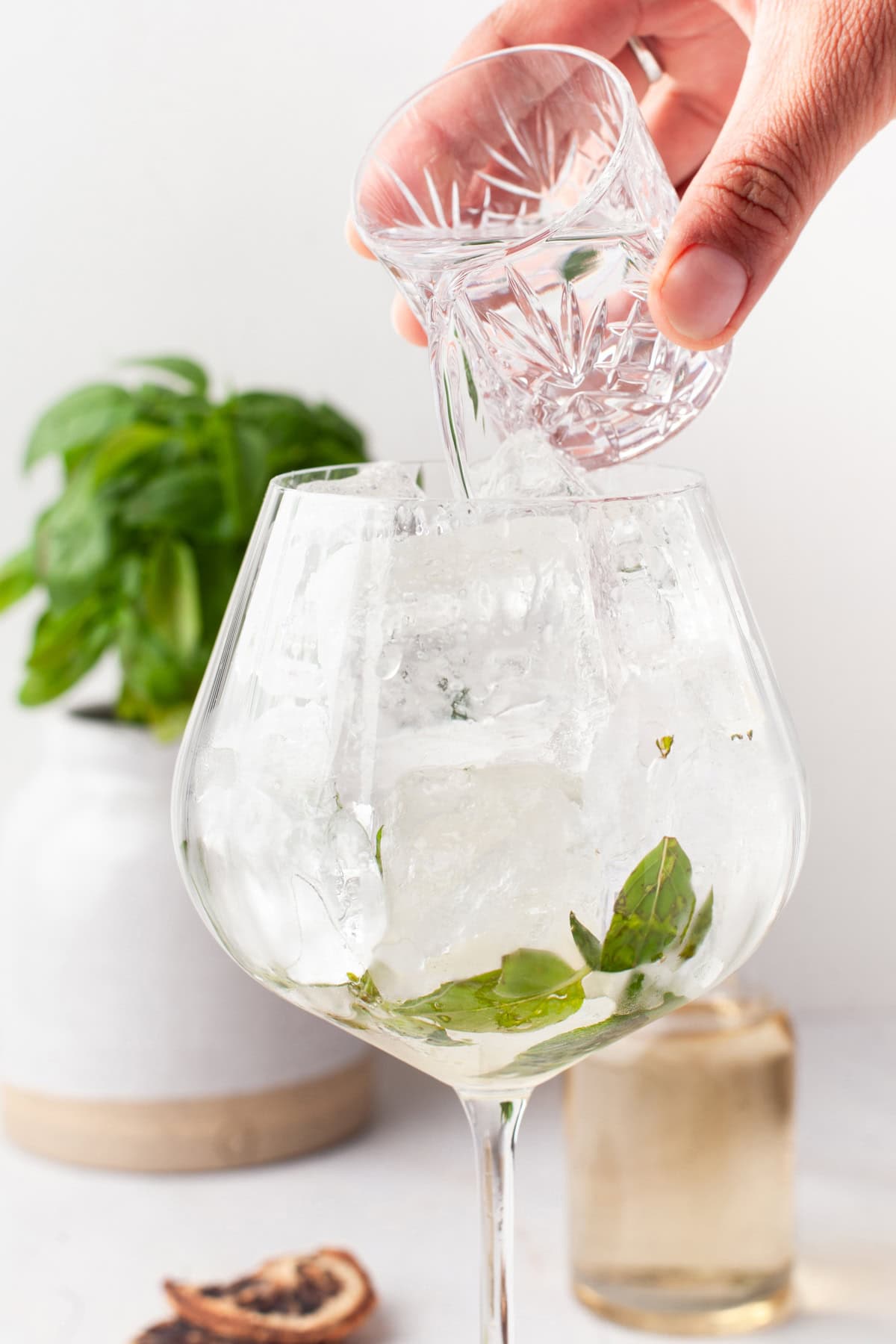 A hand pours clear liquid from a small glass into a large glass filled with ice cubes and fresh basil leaves, with a basil plant and a bottle blurred in the background.