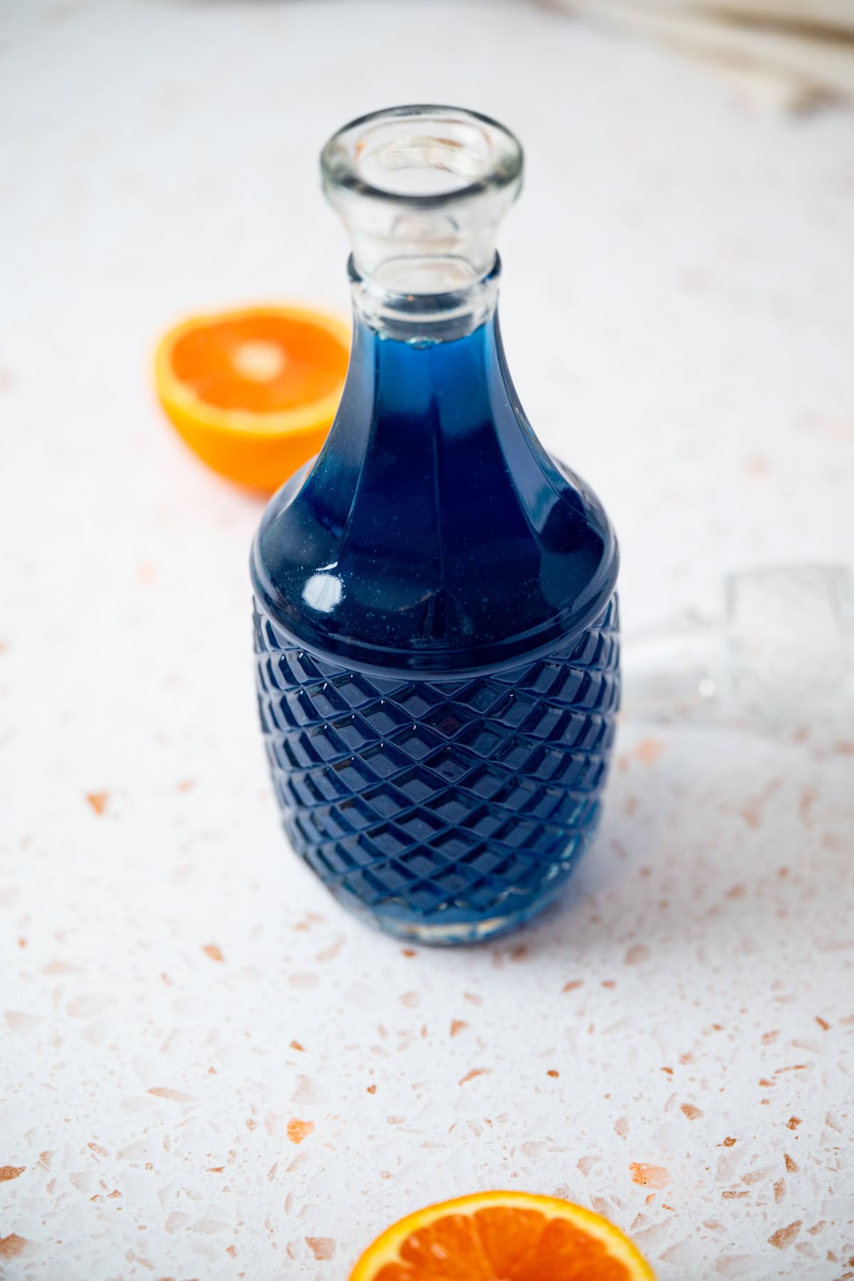 A glass decanter filled with blue liquid sits on a light, speckled countertop. Two orange slices are positioned nearby, adding a pop of color to the scene.