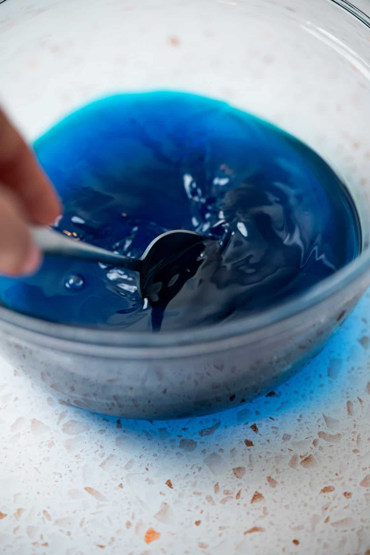A hand stirs a bowl of vibrant blue liquid with a spoon on a white countertop.