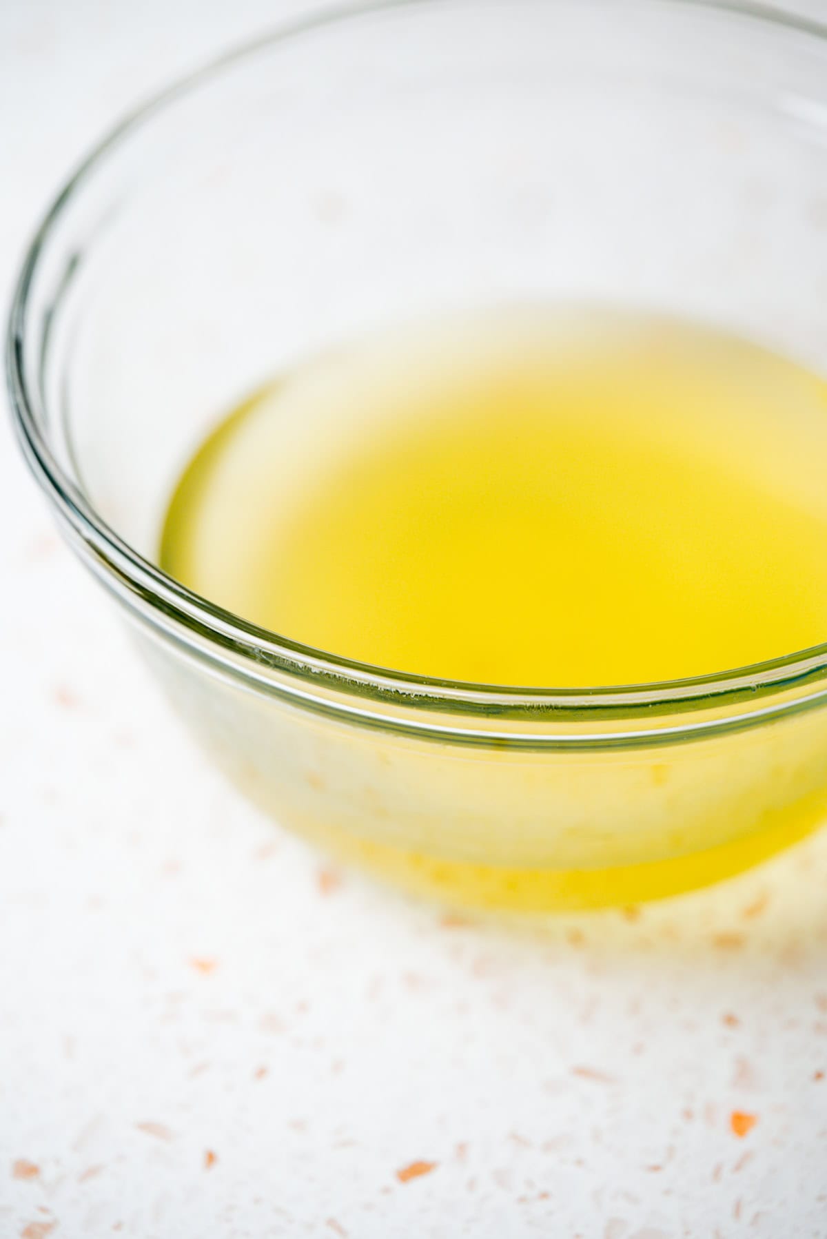 A clear glass bowl filled with a yellow liquid, possibly melted butter or oil, sits on a white countertop with small orange specks.