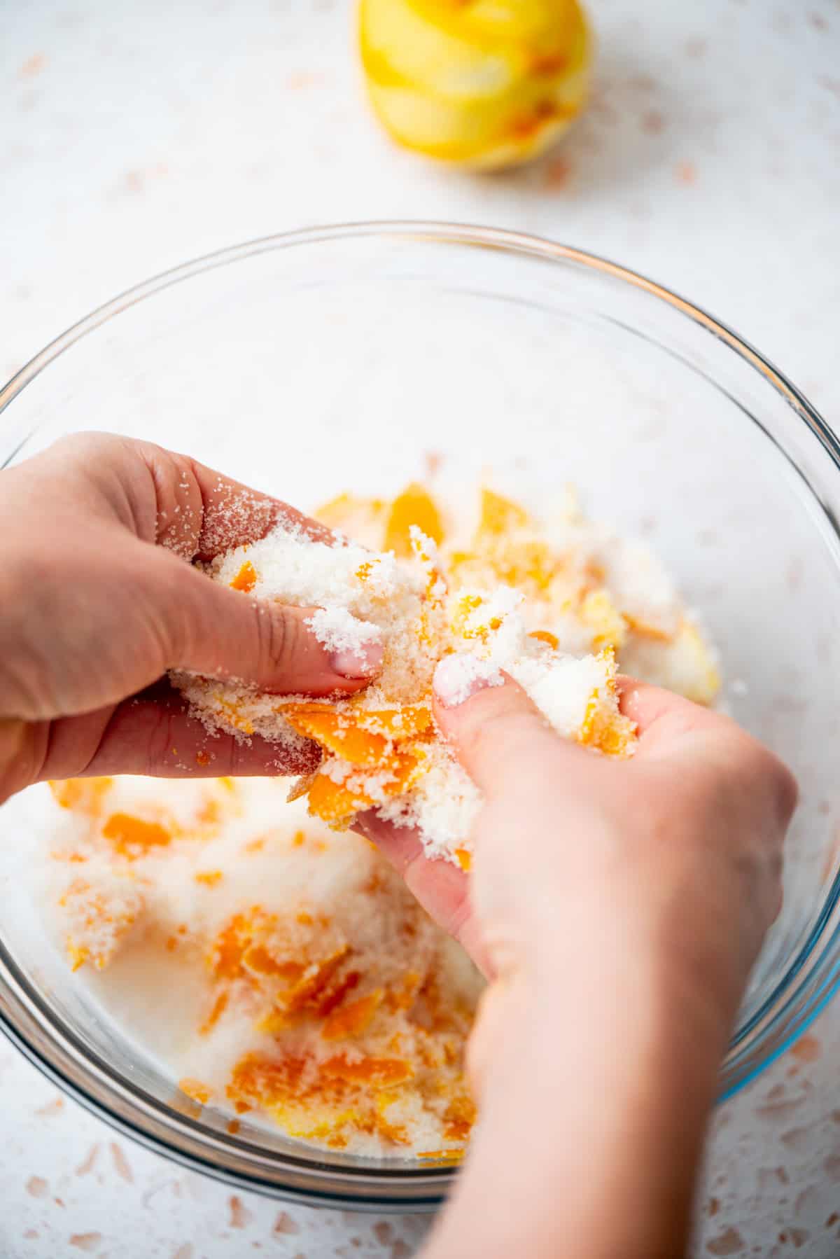 Close-up of hands mixing orange zest with sugar in a glass bowl on a light countertop; a zested orange is visible in the background.