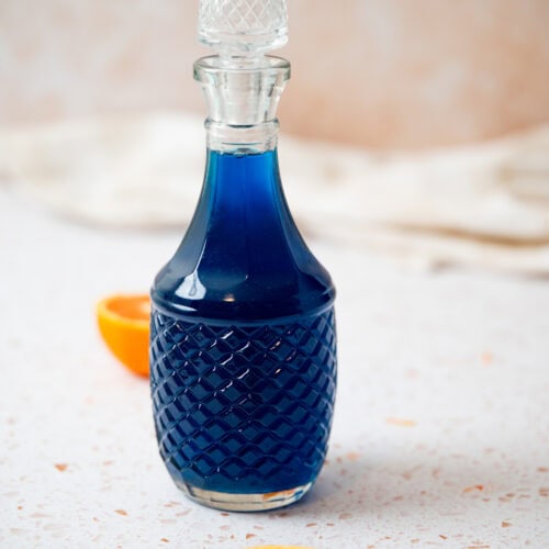 A glass decanter filled with blue liquid stands on a light speckled surface, accompanied by two halves of an orange in the foreground. The background is softly blurred.