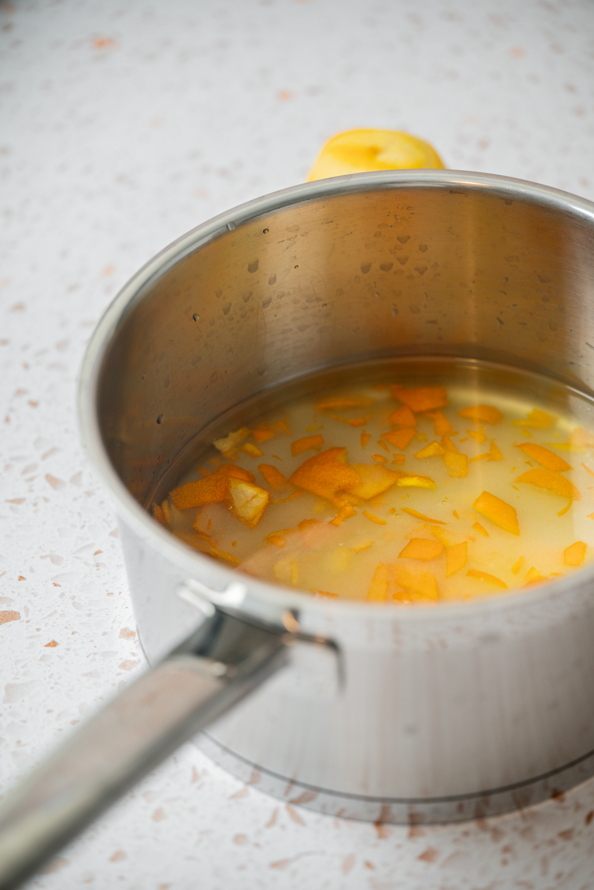 A stainless steel pot filled with water, orange peel pieces, and a cut lemon sits on a speckled countertop.