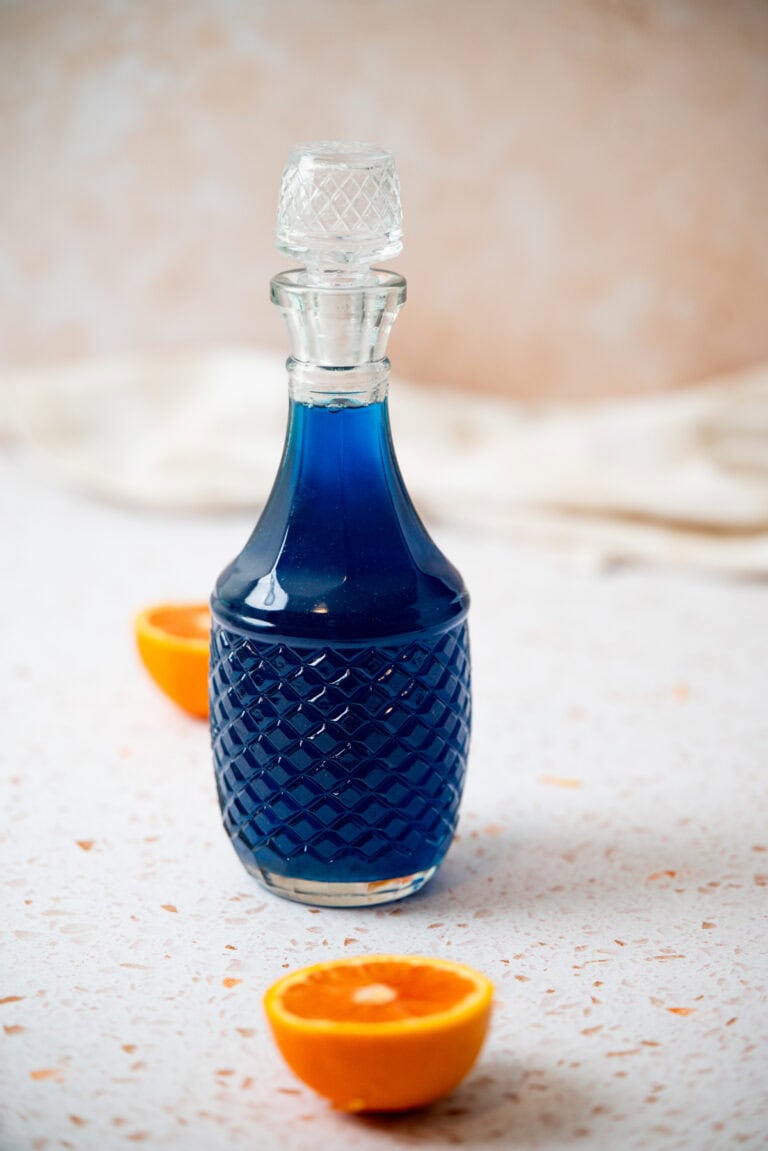 A glass decanter filled with blue liquid stands on a light speckled surface, accompanied by two halves of an orange in the foreground. The background is softly blurred.