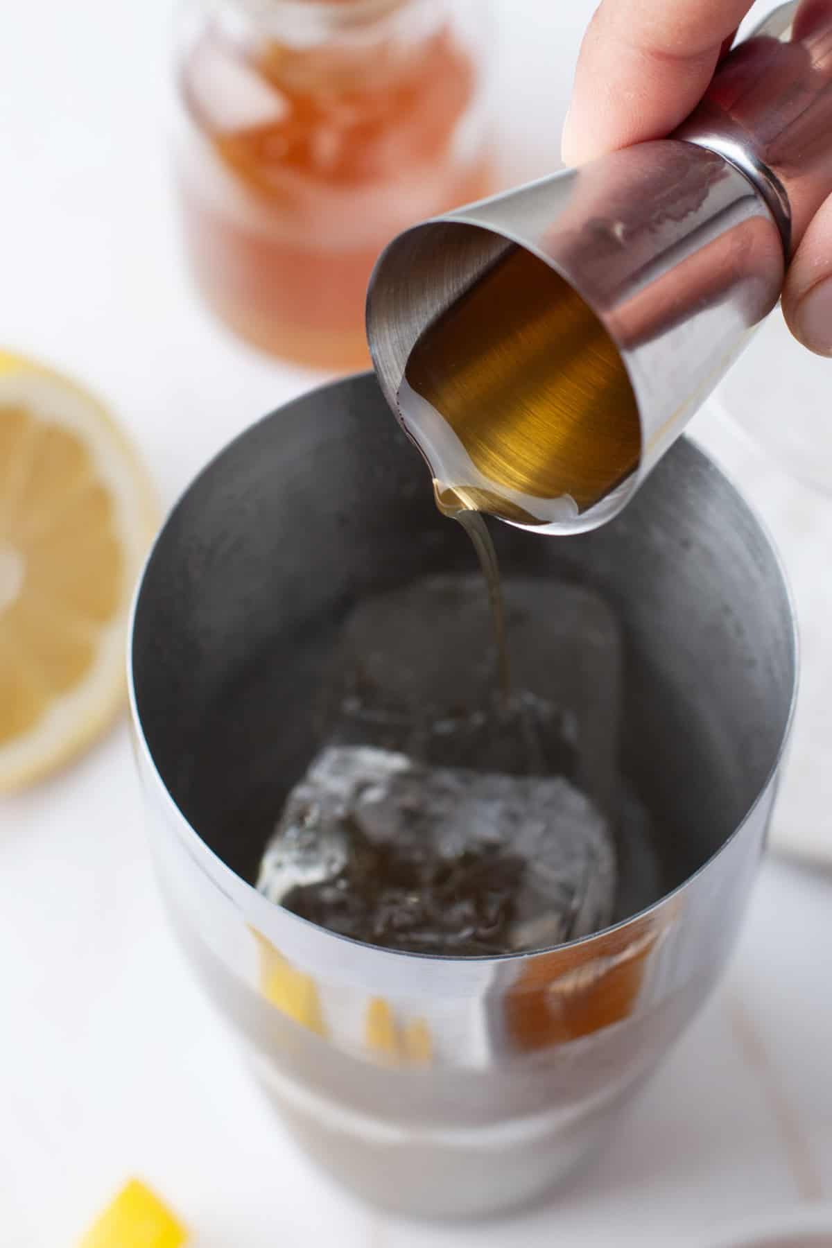 A hand pours honey from a metal jigger into a cocktail shaker with ice. A lemon slice and a jar of honey are visible in the background.