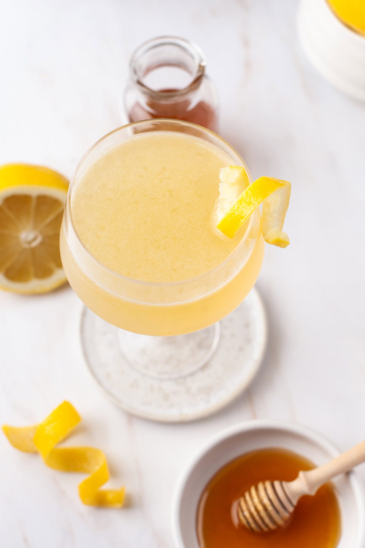 A light yellow bees knees cocktail garnished with a lemon twist sits in a coupe glass. Nearby are a halved lemon, a honey dipper in a small bowl of honey, a glass bottle of syrup, and another lemon twist on a white surface.