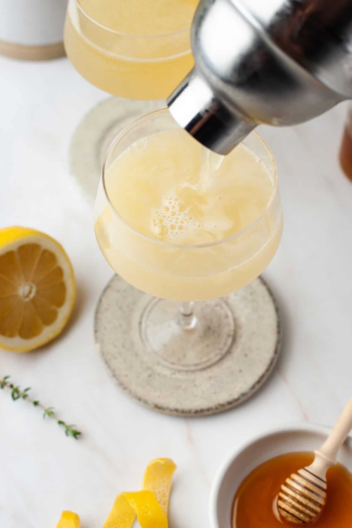 A cocktail is being poured from a shaker into a coupe glass on a coaster, with a halved lemon, honey dipper in a bowl of honey, lemon peel, and a sprig of thyme nearby on a white surface.