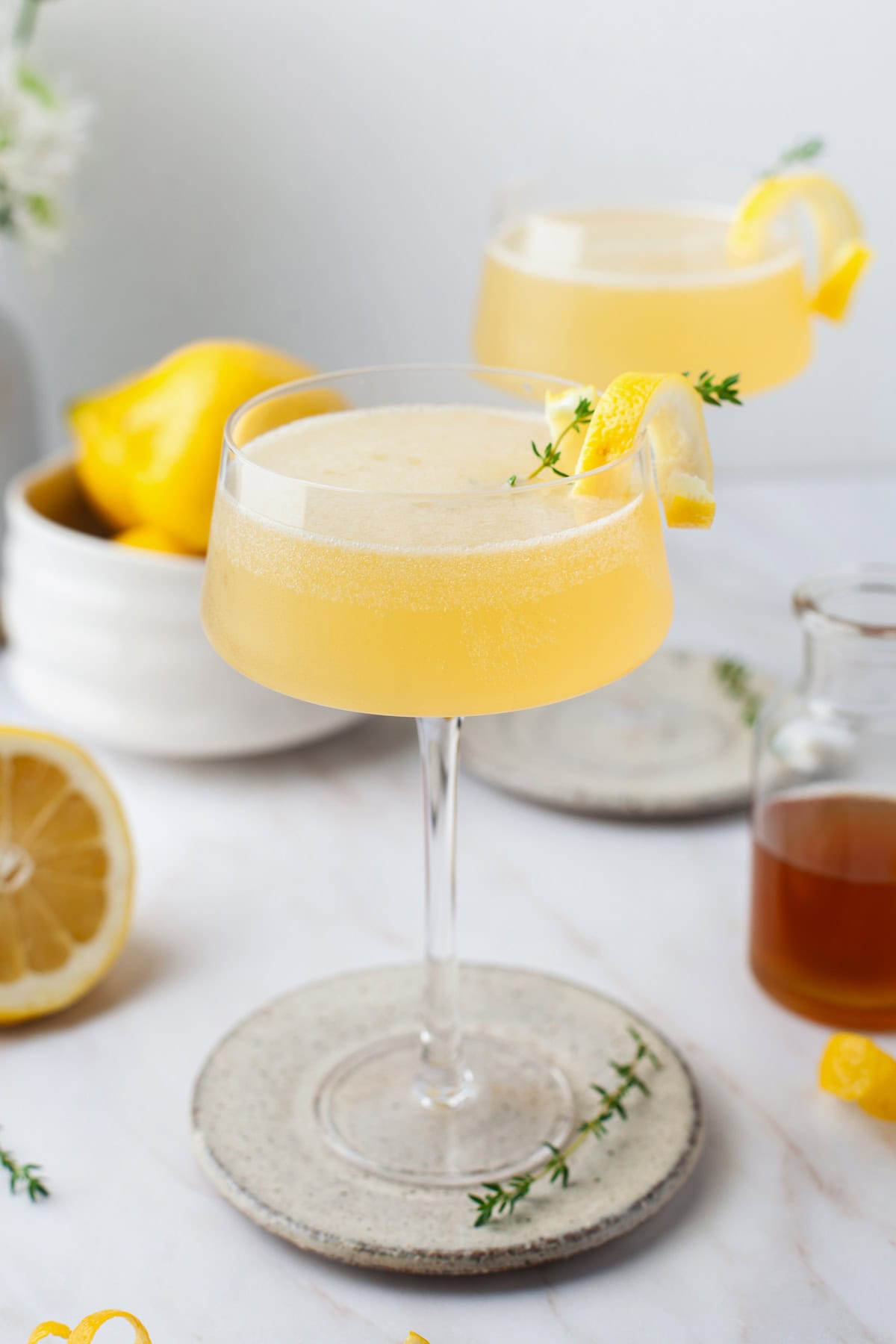 A pale yellow bees knees cocktail garnished with a lemon twist and thyme sprig sits in a coupe glass on a ceramic coaster, with lemons and a jar of honey in the background.