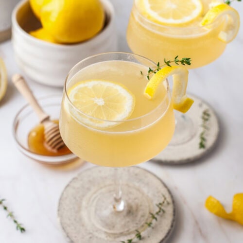 Two glasses of a light yellow drink garnished with lemon slices and thyme sprigs sit on coasters. Nearby are a bowl of lemons, a honey dipper in a small dish, and lemon peels on a white marble surface.
