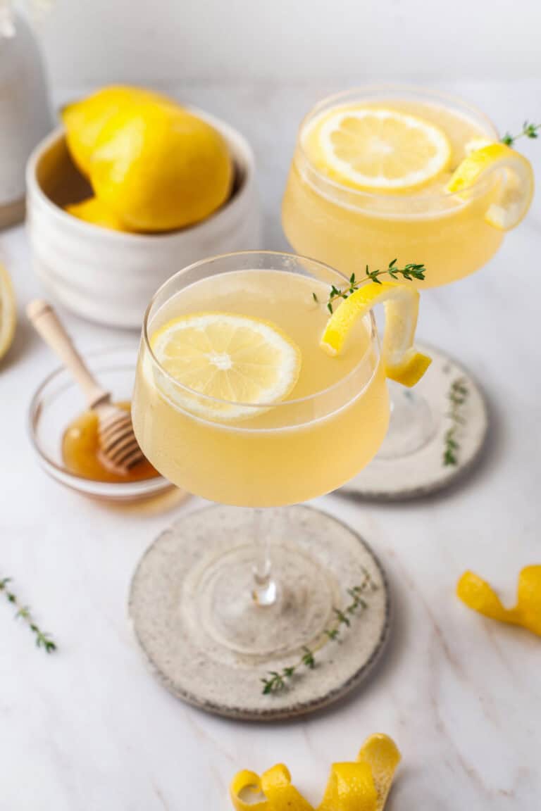 Two glasses of a light yellow drink garnished with lemon slices and thyme sprigs sit on coasters. Nearby are a bowl of lemons, a honey dipper in a small dish, and lemon peels on a white marble surface.