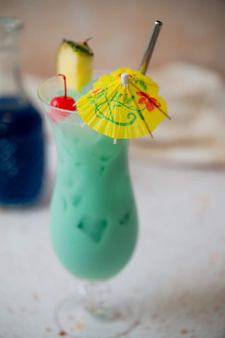A tall glass of blue-green tropical cocktail with ice, garnished with a pineapple slice, cherry, and a yellow paper umbrella, with a metal straw. A bottle with blue liquid is blurred in the background.