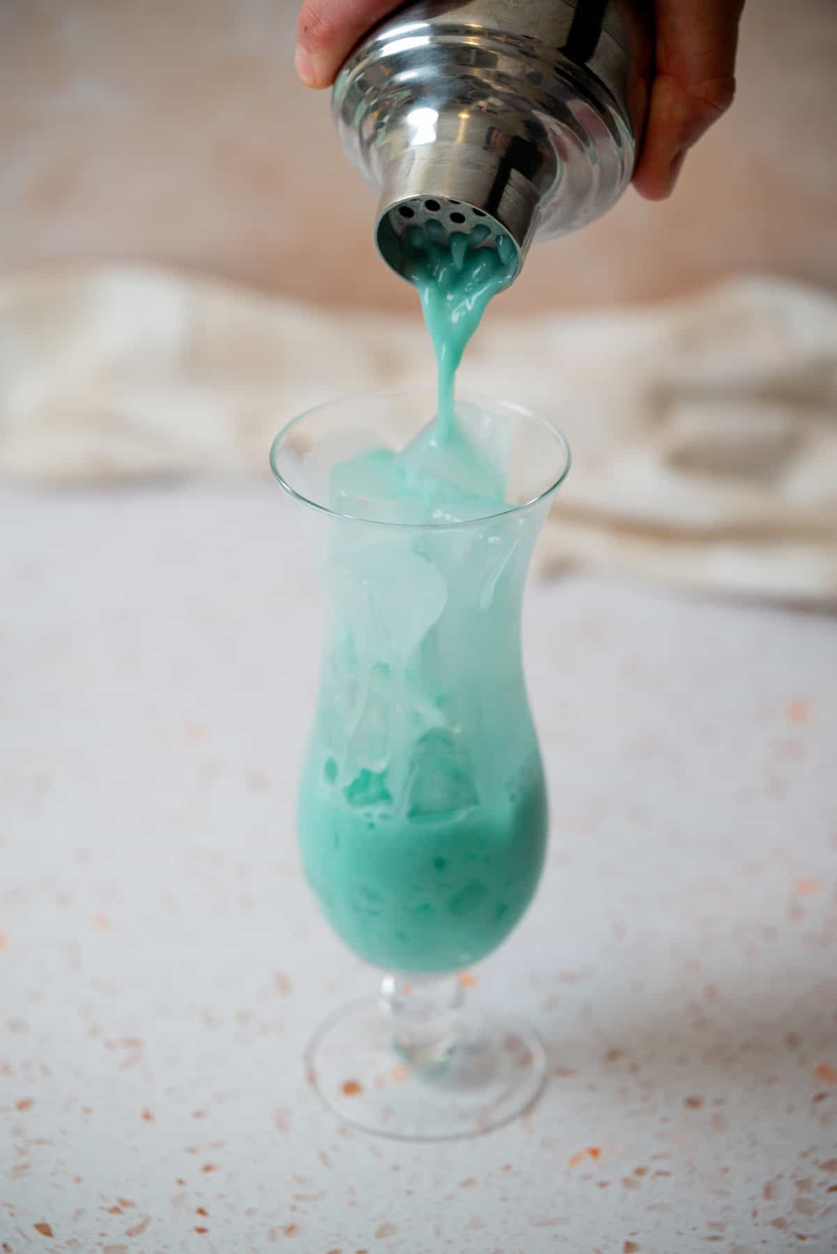 A hand pours a vibrant blue-green cocktail from a shaker into a glass filled with ice, set on a light speckled surface with a blurred white cloth in the background.