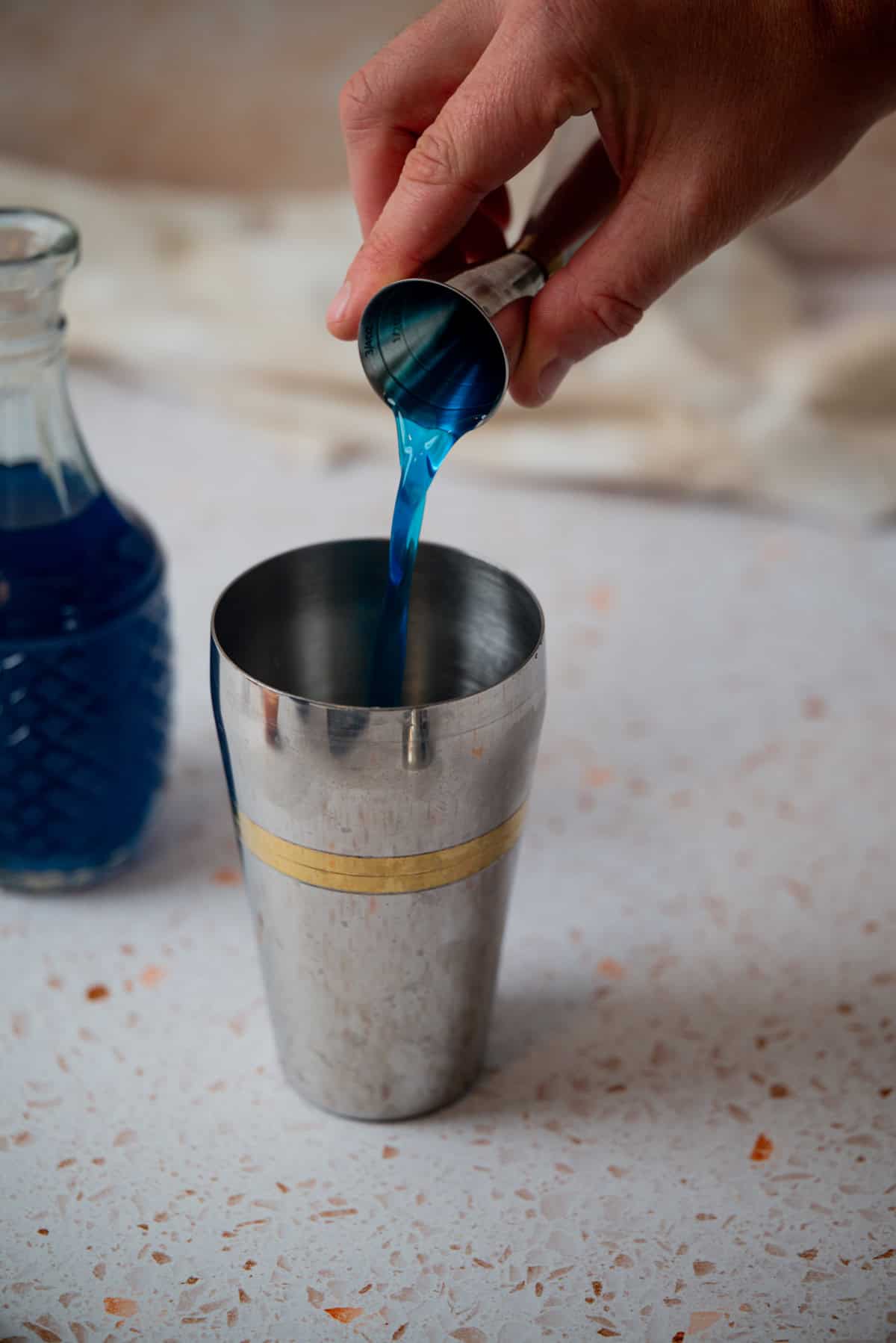 A hand pours vibrant blue liquid from a small jigger into a metal cocktail shaker; a glass bottle with more blue liquid sits nearby on a speckled countertop.