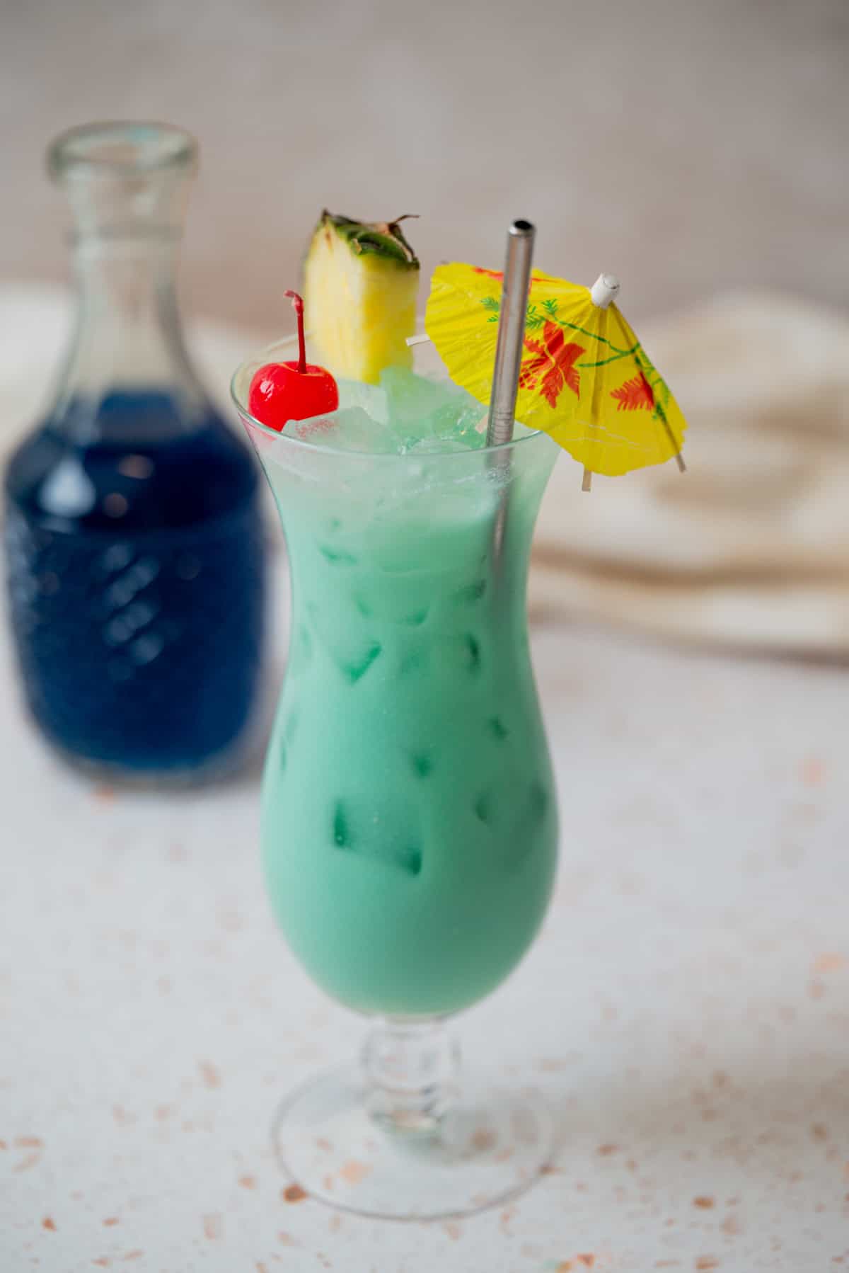 A tall glass of blue-green tropical cocktail with ice, topped with a pineapple wedge, maraschino cherry, and a yellow paper umbrella, sits on a light surface. A small bottle of dark blue liquid is in the background.