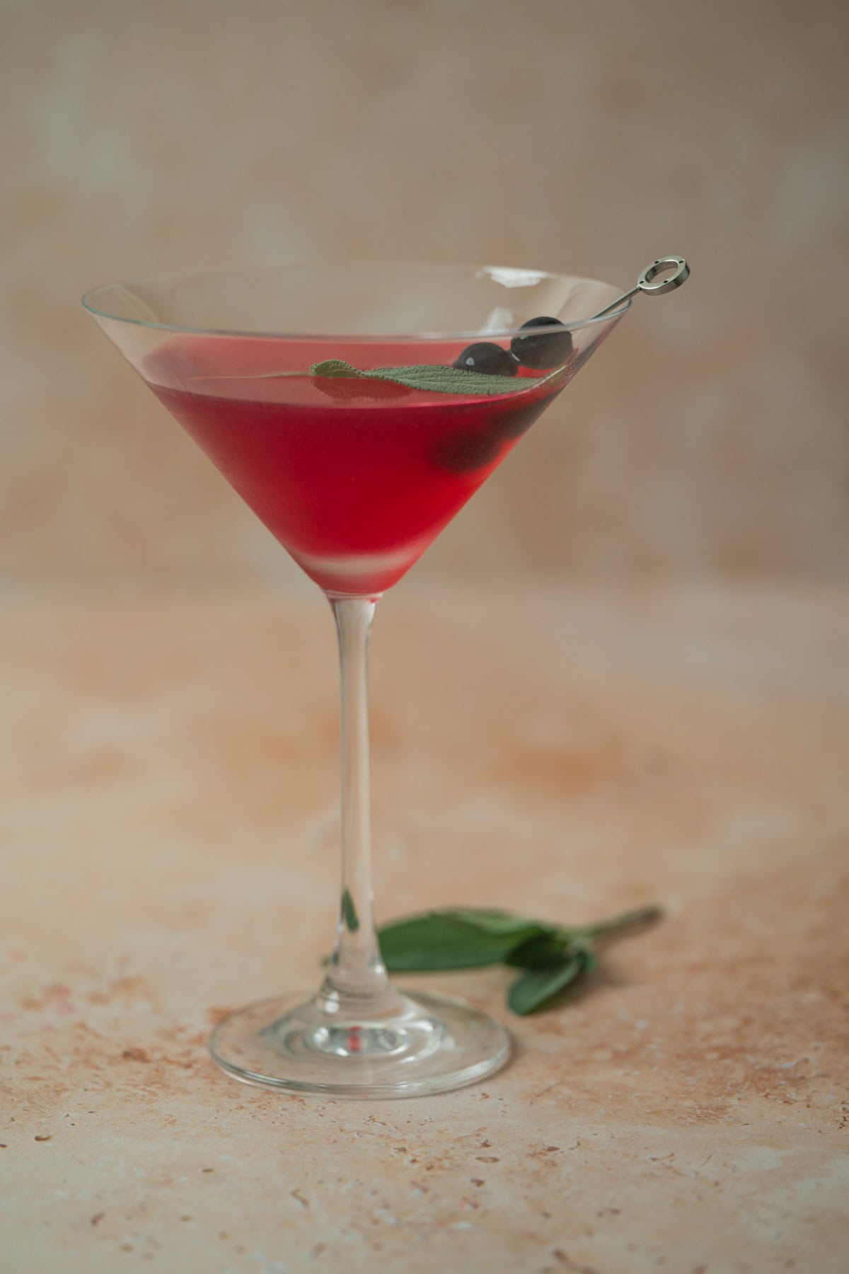 A martini glass filled with a blueberry sage martini garnished with two blueberries and a green leaf on a cocktail pick, sits on a light textured surface with extra leaves nearby.