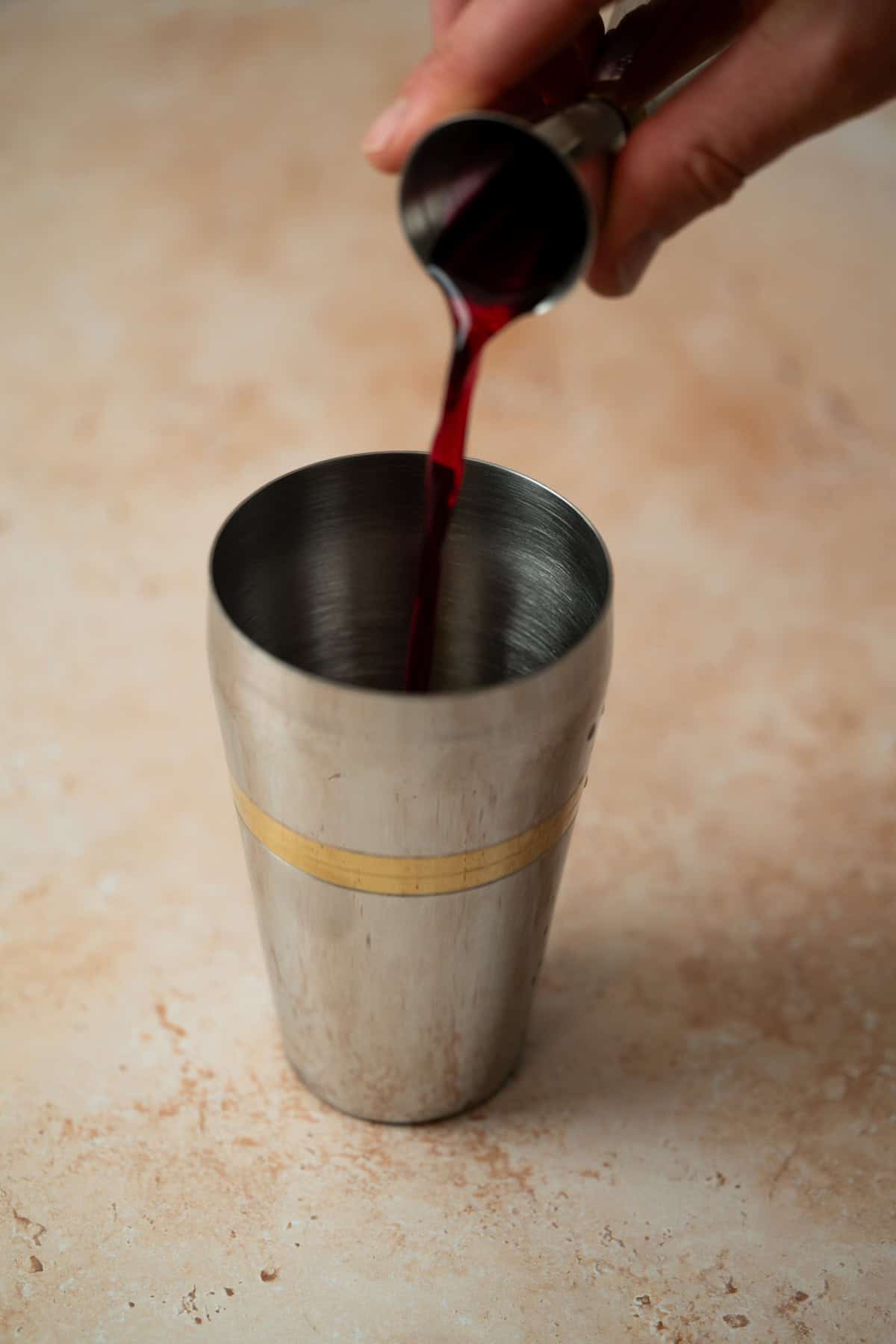 A hand pours a deep red liquid from a jigger into a stainless steel cocktail shaker on a light beige surface.