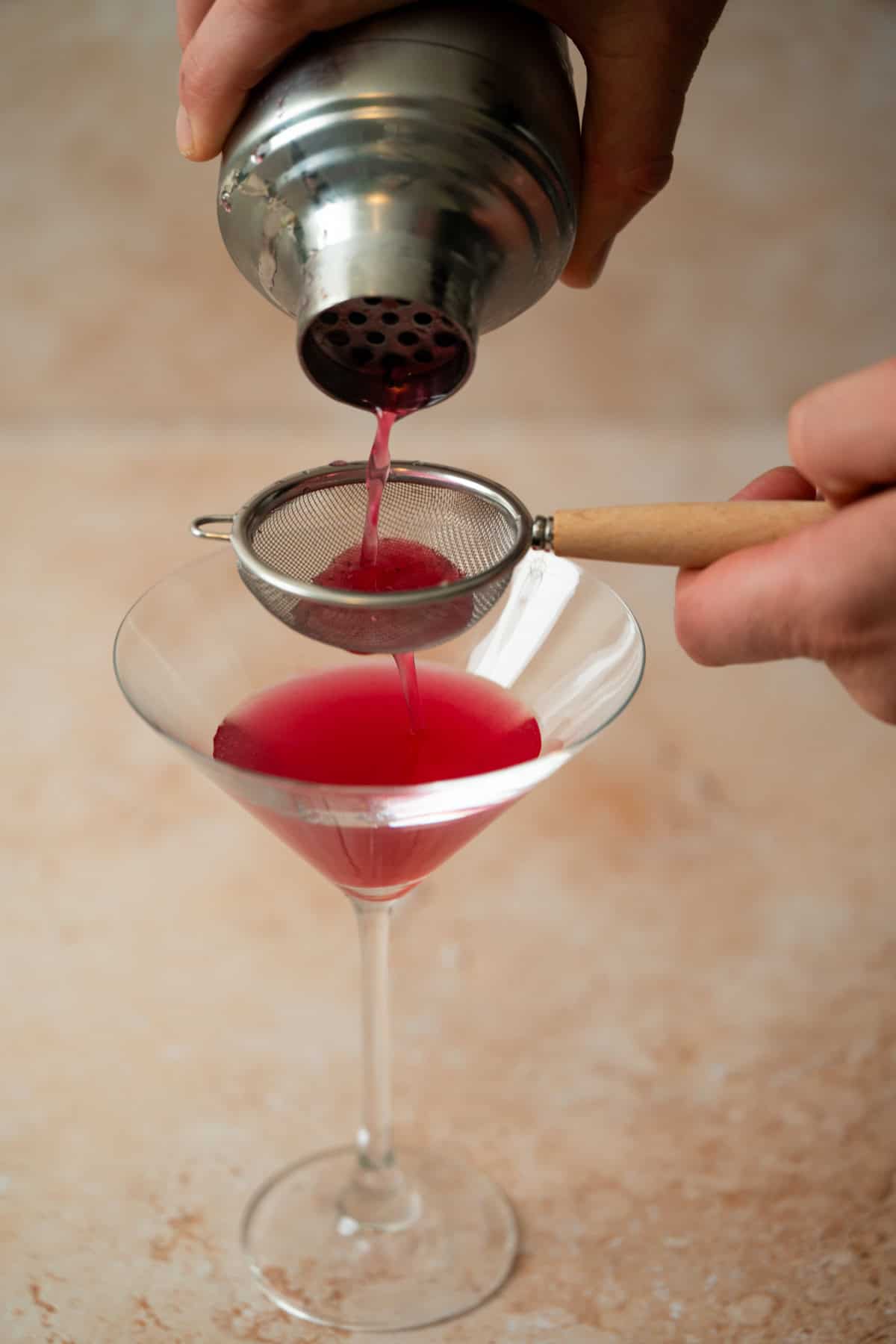 A person pours a bright pink cocktail from a shaker through a handheld strainer into a martini glass on a light-colored surface.