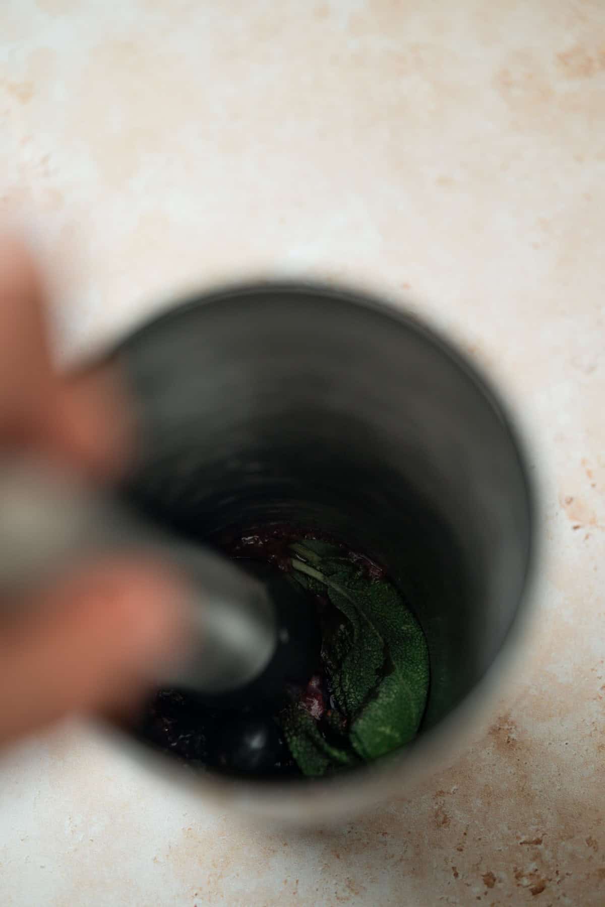 A close-up image of a metal mortar and pestle in use, with green and purple ingredients being crushed inside the mortar, viewed from above on a light-colored surface.