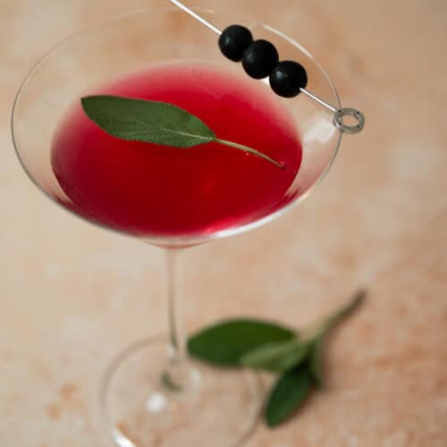 A martini glass filled with blueberry sage martini garnished with a fresh sage leaf and three black olives skewered on a cocktail pick, sits on a light, textured surface with sage leaves nearby.