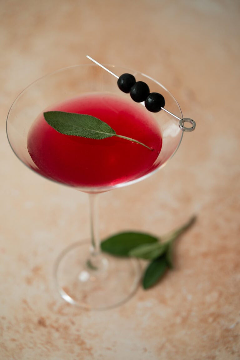 A martini glass filled with blueberry sage martini garnished with a fresh sage leaf and three black olives skewered on a cocktail pick, sits on a light, textured surface with sage leaves nearby.