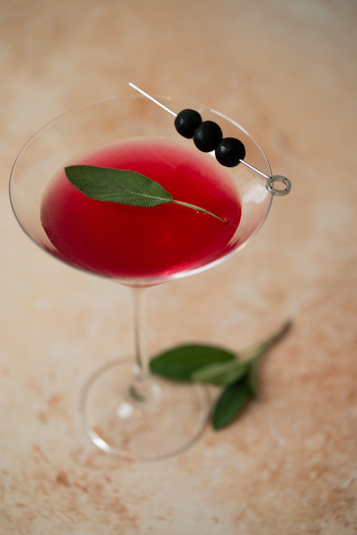 A martini glass filled with blueberry sage martini garnished with a fresh sage leaf and three black olives skewered on a cocktail pick, sits on a light, textured surface with sage leaves nearby.