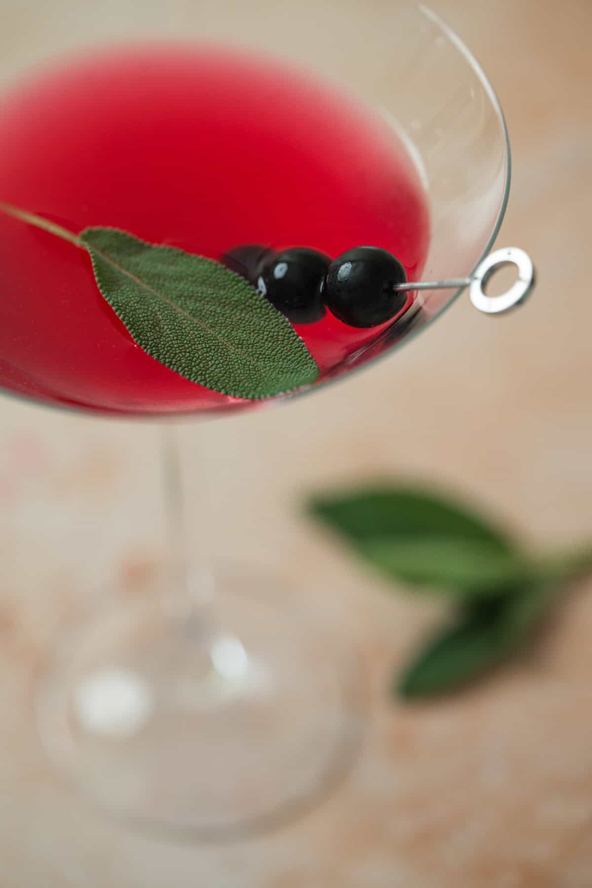 A vibrant red blueberry sage martini in a martini glass, garnished with blueberries and a fresh green sage leaf on a metal pick. An extra sage leaf lies blurred in the background.