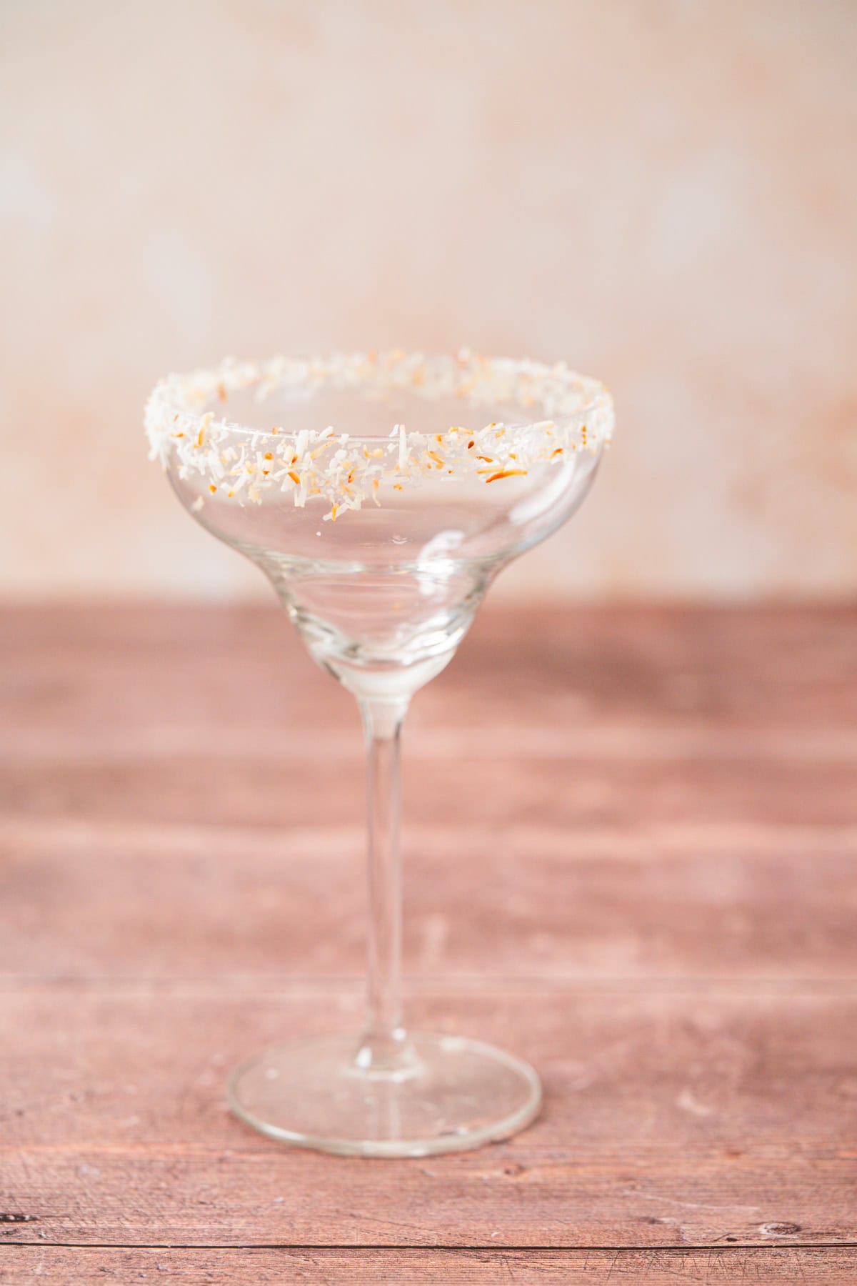 A margarita glass with a rim coated in shredded coconut sits on a wooden surface against a soft, beige background. The glass is empty.