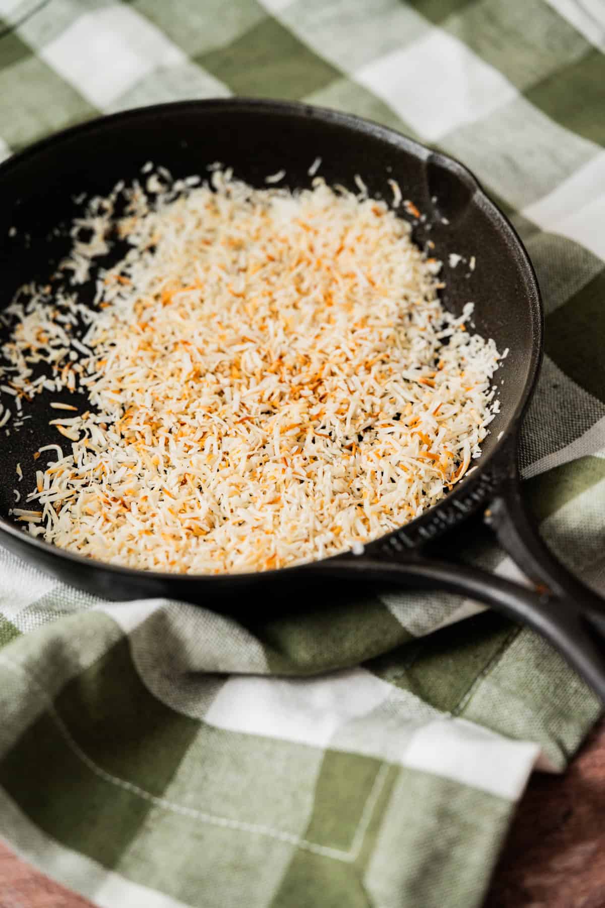 A black skillet with toasted shredded coconut sits on a green and white checkered cloth.