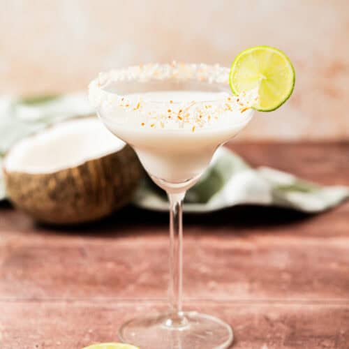 A creamy white cocktail in a margarita glass with a toasted coconut rim, garnished with a lime wheel. Slices of lime and a halved coconut are nearby on a wooden surface.
