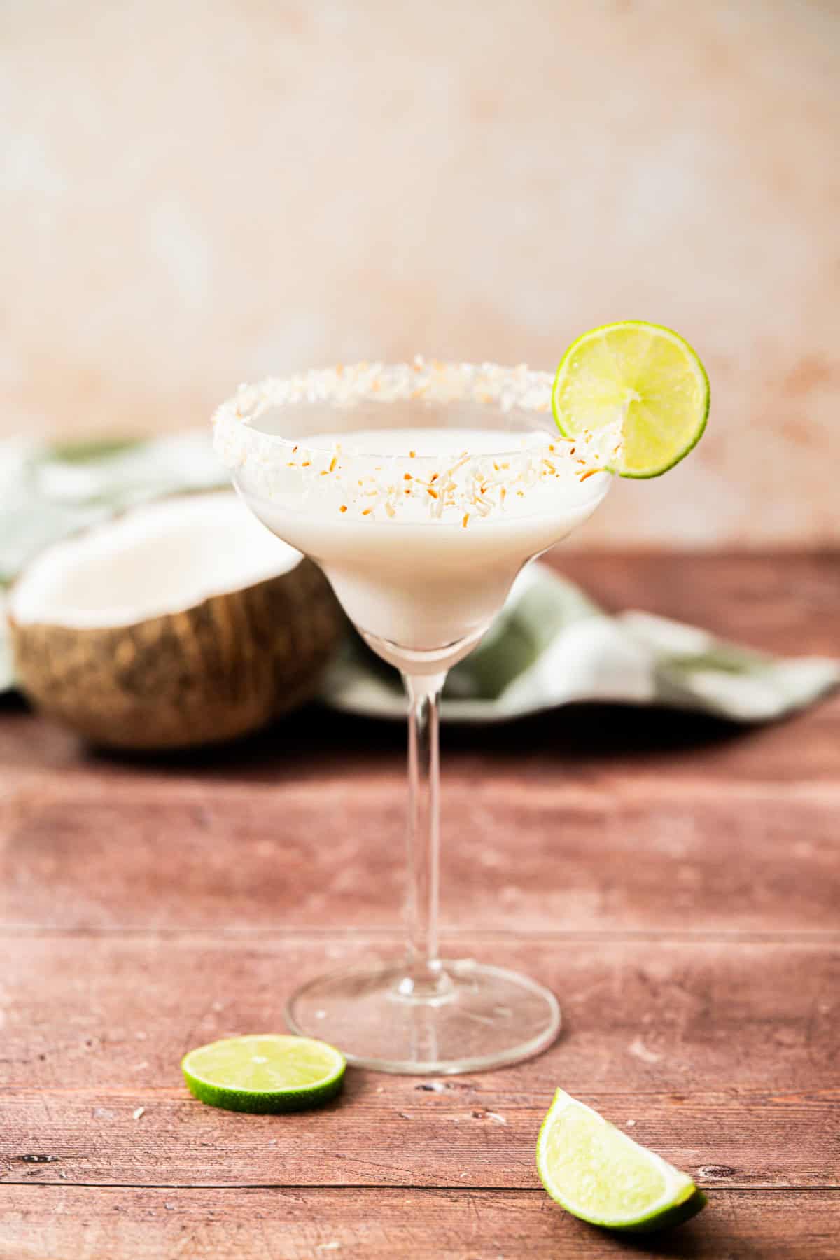 A creamy white cocktail in a margarita glass with a toasted coconut rim, garnished with a lime wheel. Slices of lime and a halved coconut are nearby on a wooden surface.
