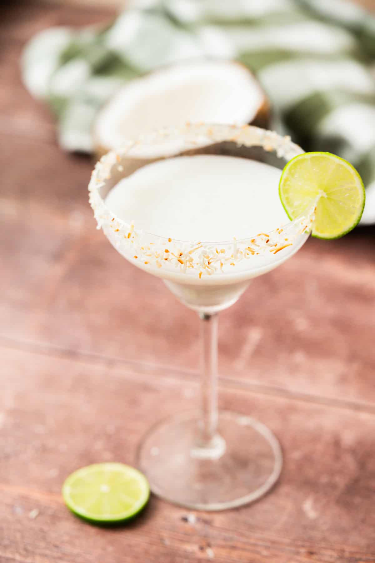 A margarita glass with a toasted coconut rim, filled with a creamy white drink, garnished with a lime wheel. A halved lime and a green-checkered cloth with a coconut are in the blurred background.
