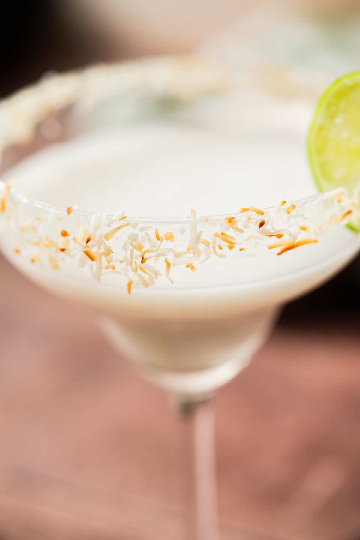 A close-up of a creamy white cocktail in a glass garnished with toasted coconut flakes on the rim and a slice of lime.