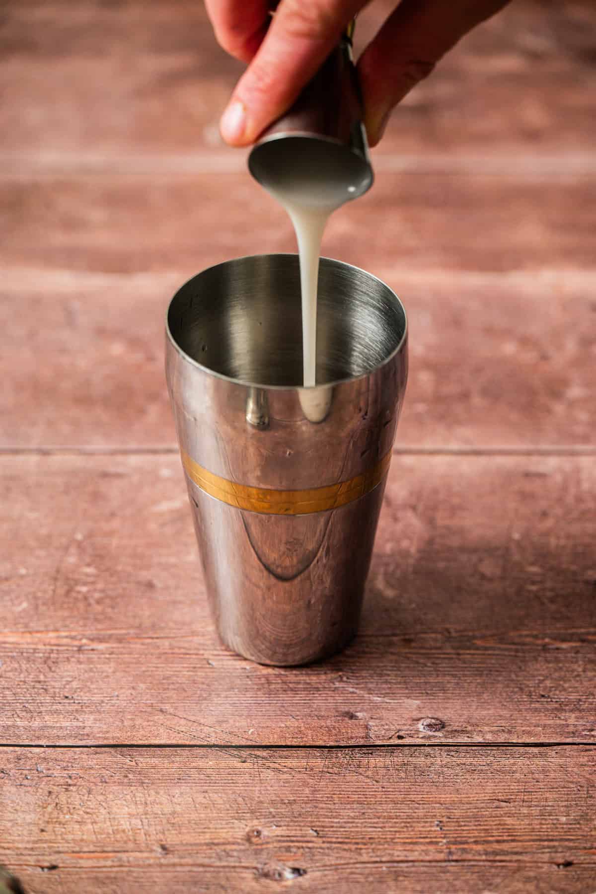 A hand pours a creamy white liquid from a small metal cup into a stainless steel cocktail shaker on a wooden surface.