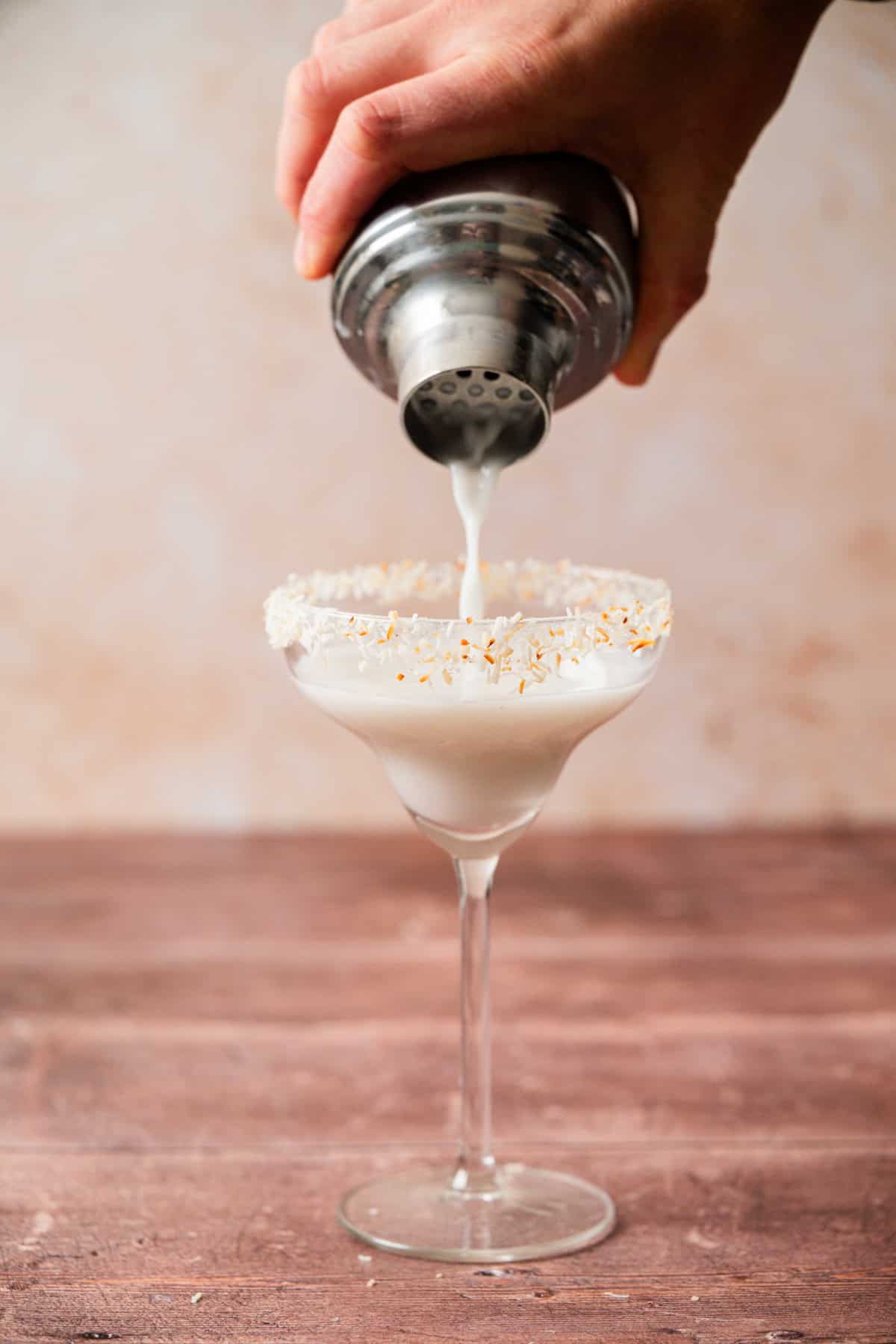 A hand pours a creamy white cocktail from a metal shaker into a margarita glass with a coconut-flake rim, set on a wooden surface.