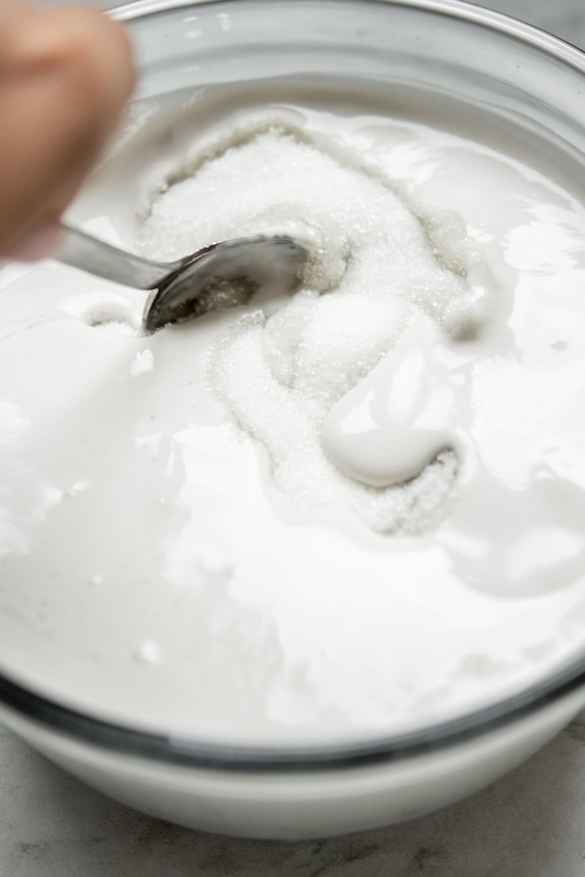 A close-up of a hand stirring white granulated sugar into a bowl of creamy liquid, possibly yogurt or a similar mixture, using a spoon.