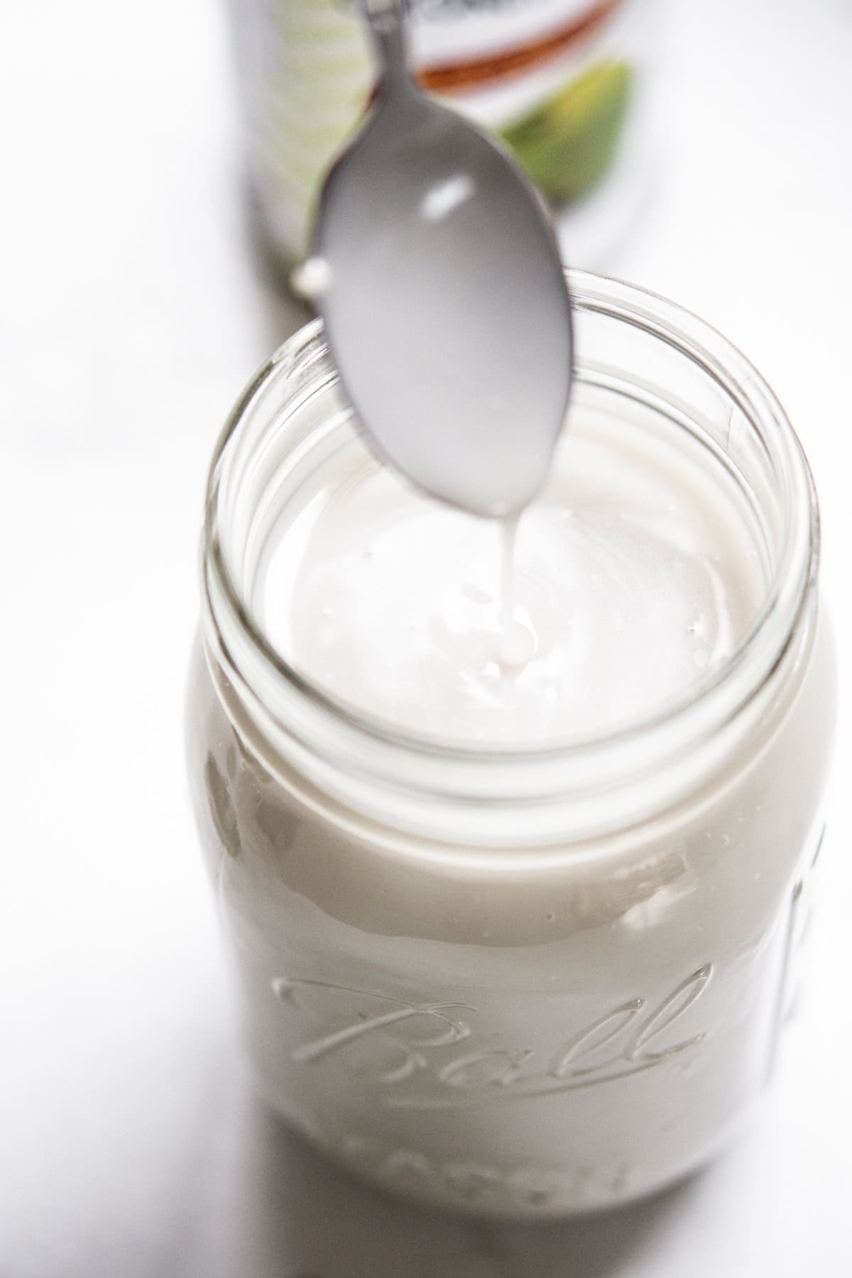 A spoon drips thick, white liquid into a glass jar filled with a creamy substance, likely yogurt or coconut milk, on a white surface.