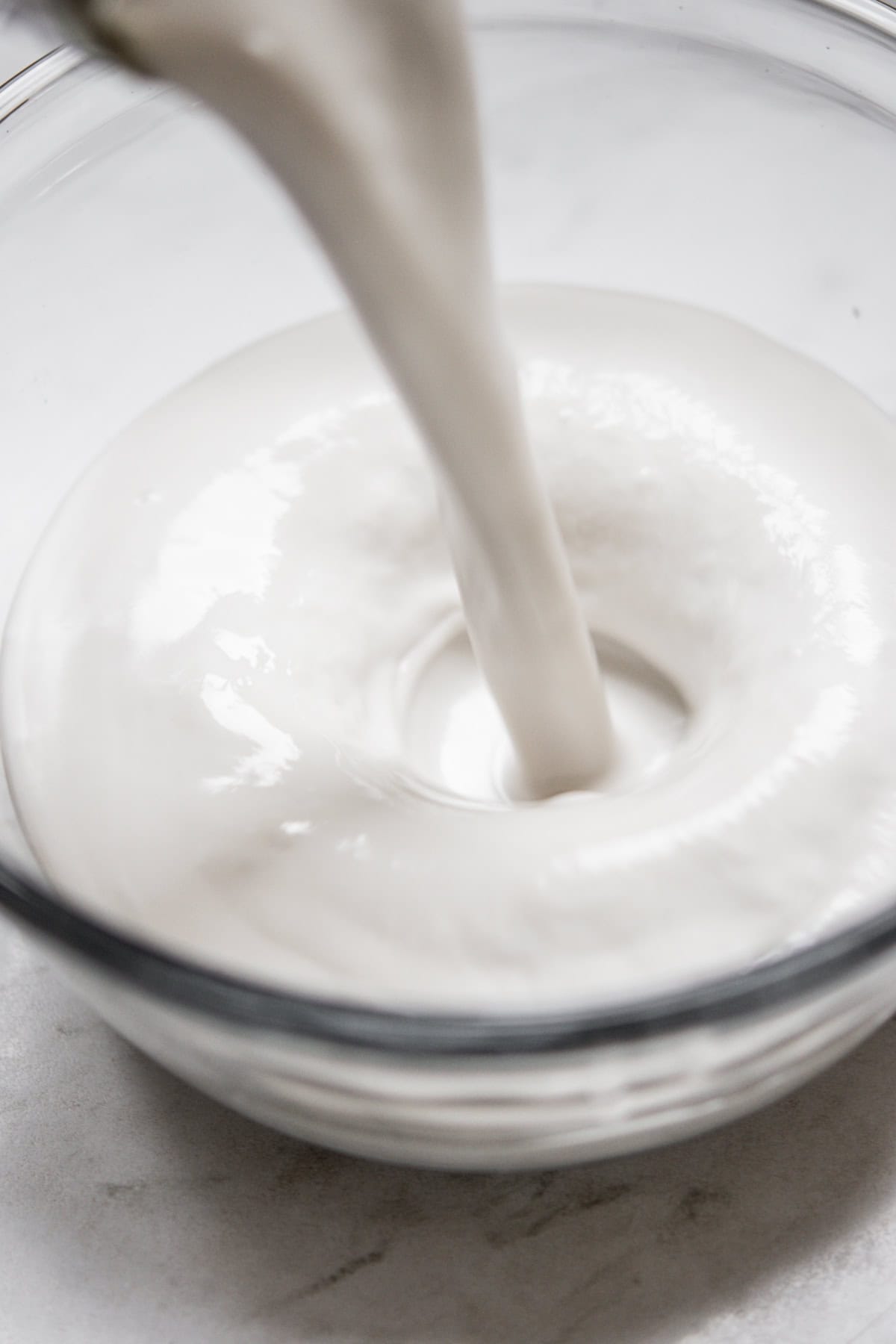 Thick white liquid being poured into a clear glass bowl, creating a smooth, creamy swirl on a light surface.