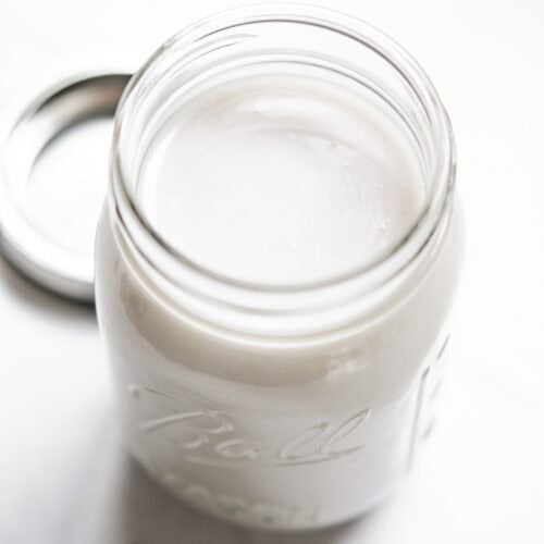 A glass Ball mason jar filled with homemade cream of coconut sits on a white surface with its lid off and placed nearby.