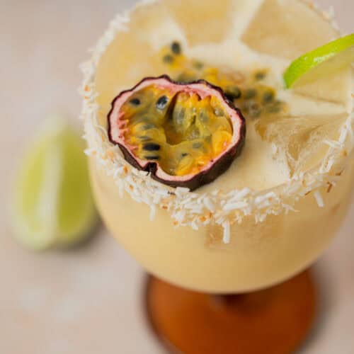 A creamy yellow passionfruit coconut daiquiri in a glass with a coconut rim, garnished with a halved passion fruit and a lime twist. Ice cubes are visible in the drink, with lime wedges blurred in the background.