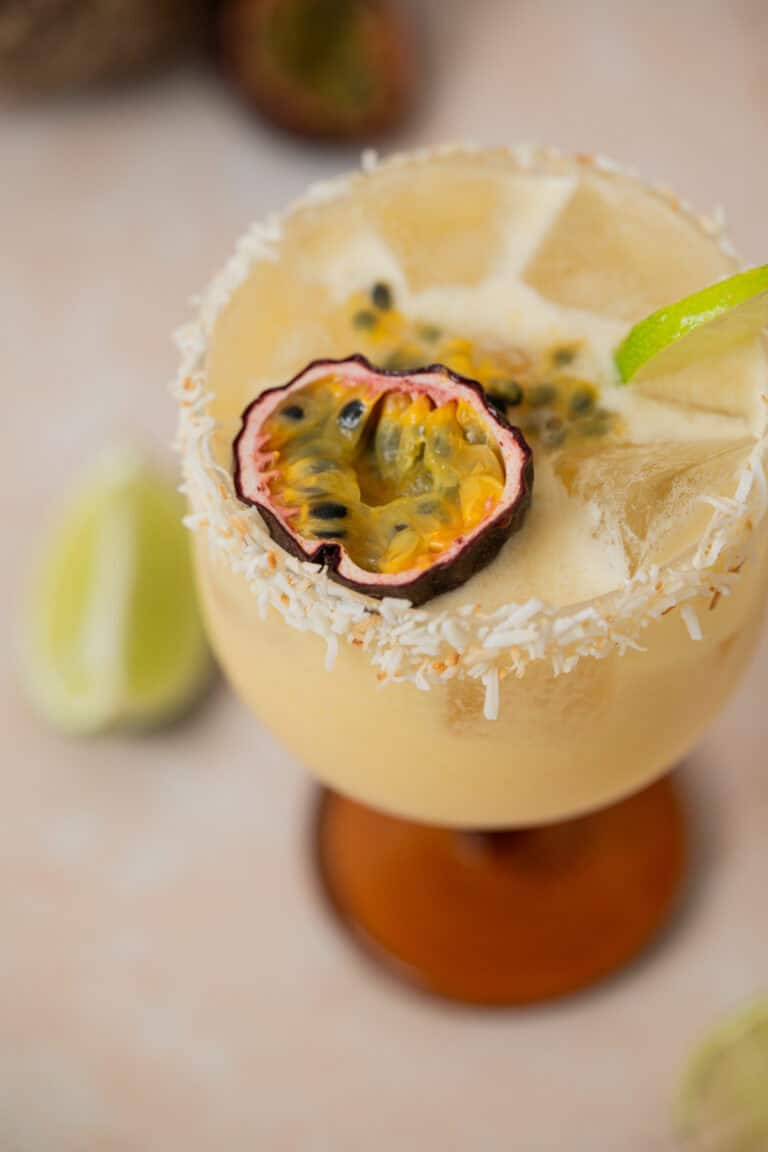 A creamy yellow passionfruit coconut daiquiri in a glass with a coconut rim, garnished with a halved passion fruit and a lime twist. Ice cubes are visible in the drink, with lime wedges blurred in the background.