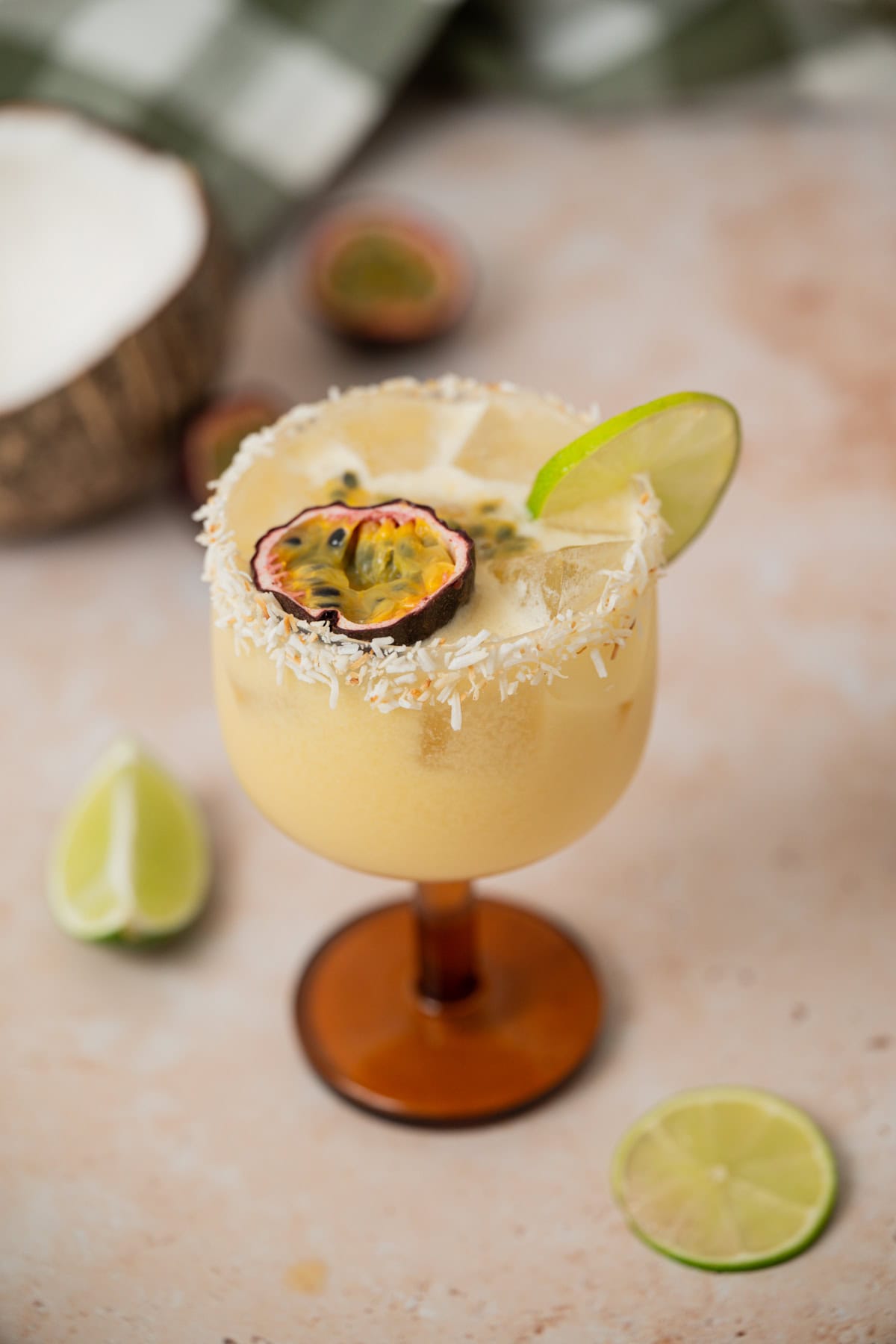 A creamy yellow cocktail in a stemmed glass, garnished with a lime wheel, half a passion fruit, and shredded coconut on the rim. Sliced lime and passion fruit are in the background on a light surface.