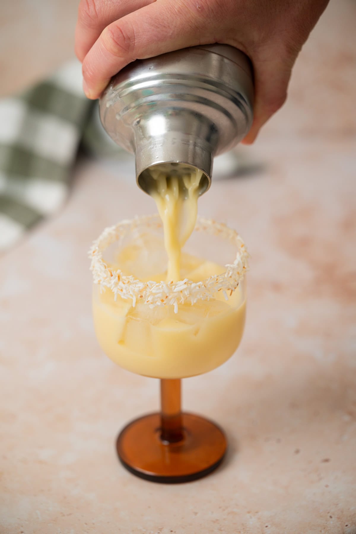 A hand pours a creamy yellow drink from a shaker into a glass with a toasted coconut rim on a light surface. A green and white checkered cloth is blurred in the background.