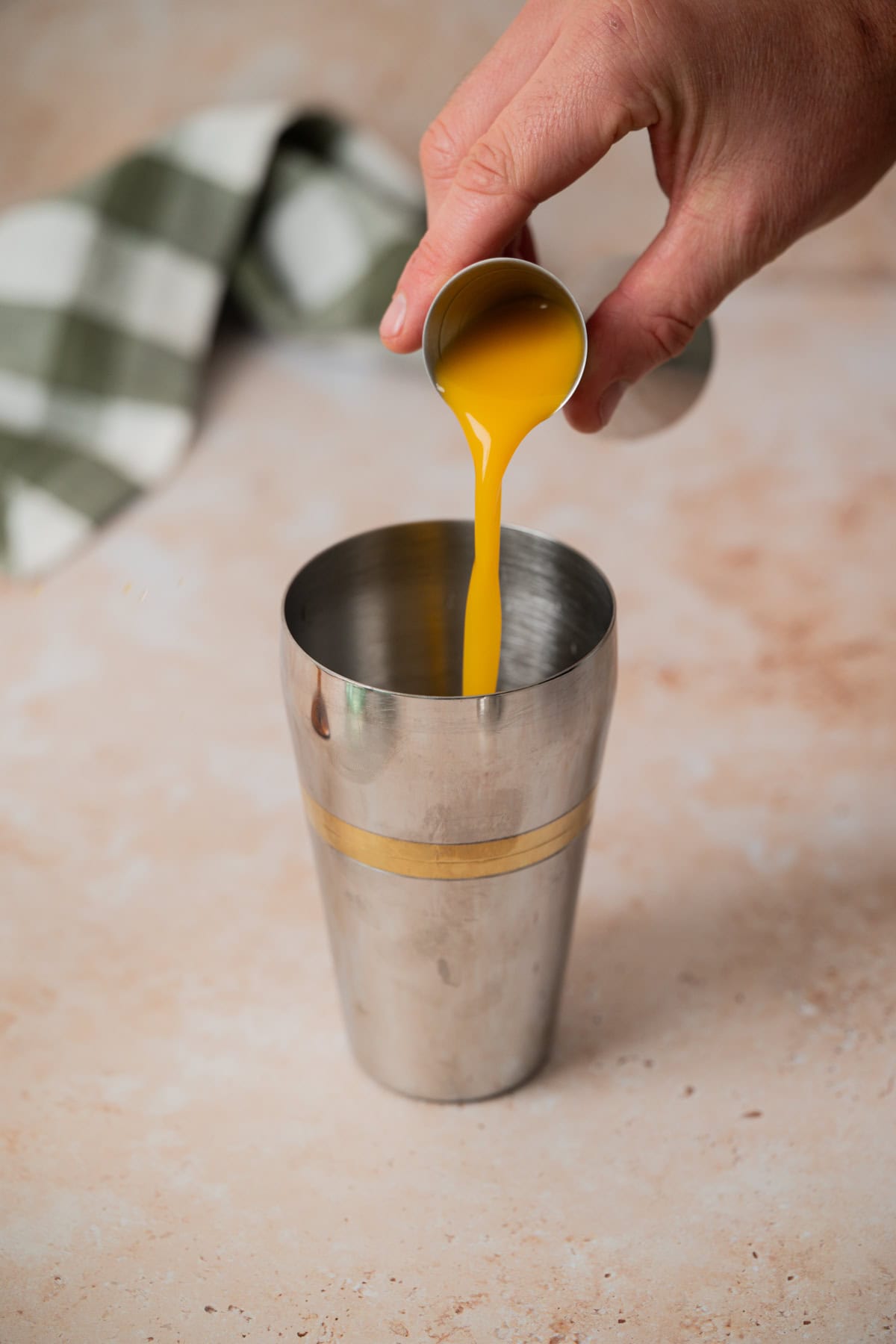 A hand pours orange juice from a small metal cup into a metal cocktail shaker, with a green and white checkered cloth blurred in the background.