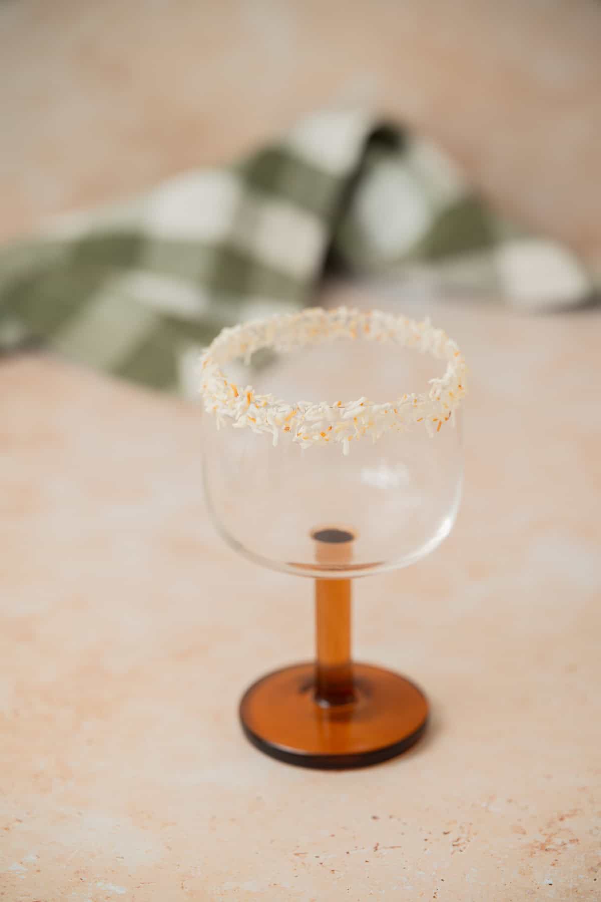 A clear glass with a brown stem sits on a light surface. The rim of the glass is coated with shredded coconut. In the blurred background, there is a green and white checkered cloth.