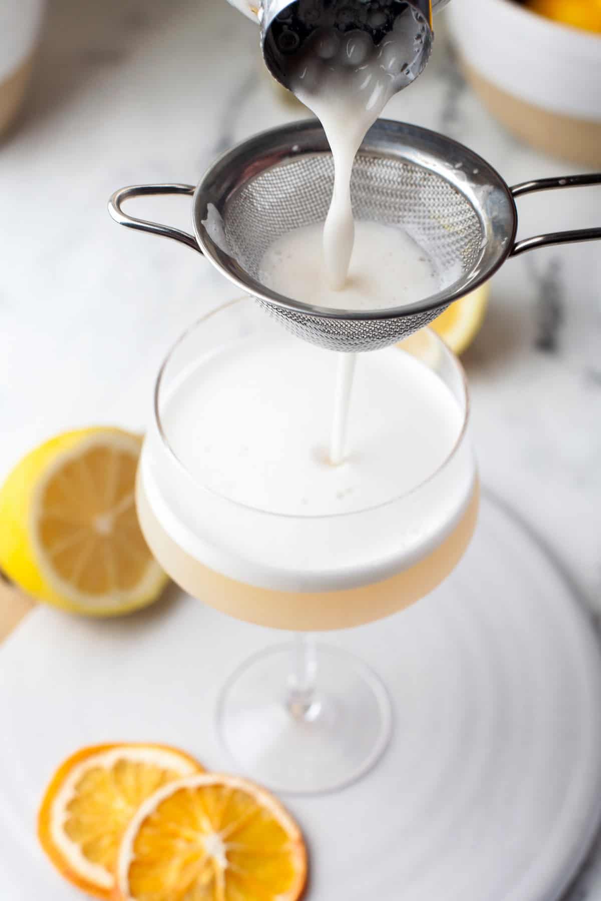 A creamy white cocktail is being poured through a mesh strainer into a coupe glass. Slices of lemon and dried orange garnish the plate beside the glass; a halved lemon is in the background.