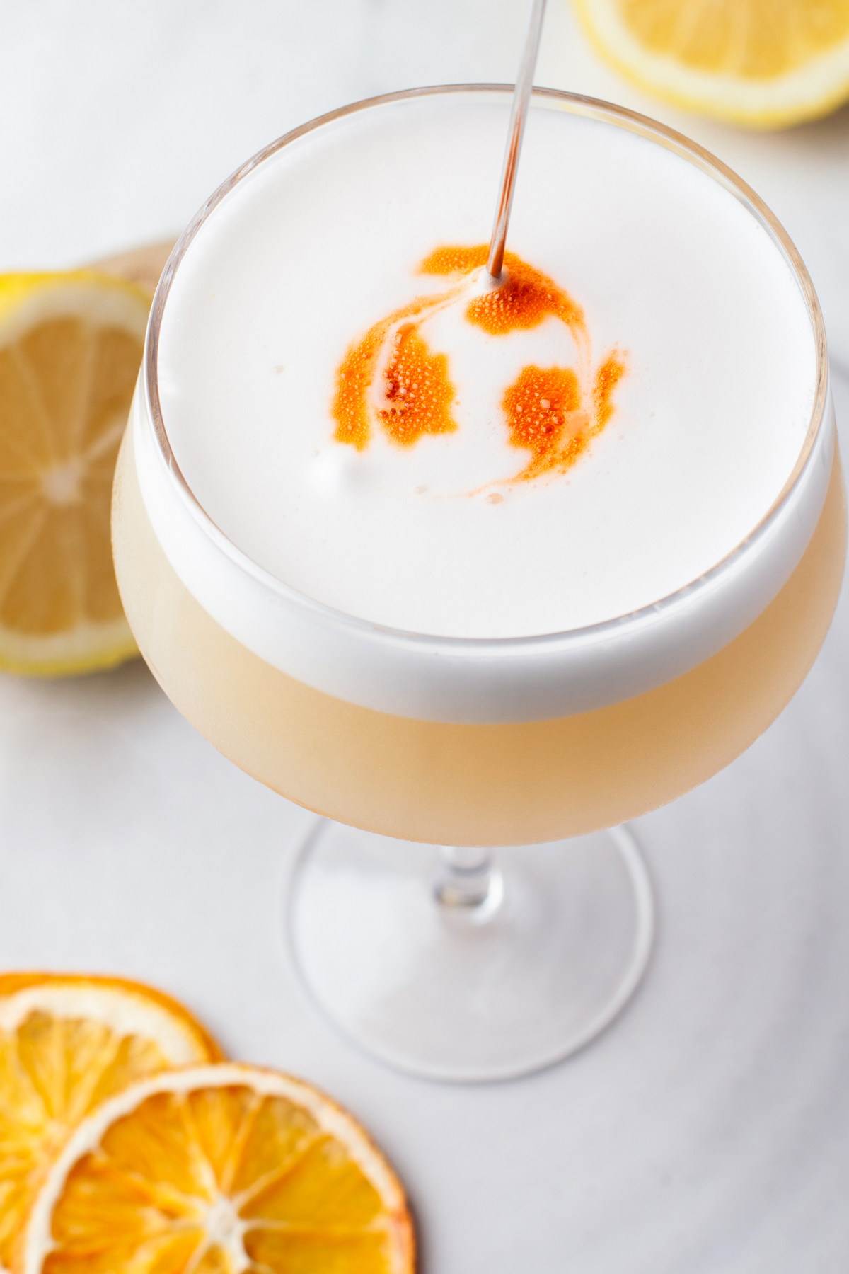 A cocktail with a frothy white top is garnished with drops of orange bitters. Slices of fresh and dried orange are placed beside the glass on a white surface.