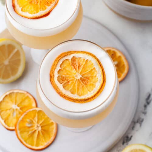 Two coupe glasses filled with a frothy rum sour cocktail, each garnished with a dried orange slice, sit on a white plate. Slices of dried and fresh lemon surround the glasses, with a bowl of fresh lemons in the background.