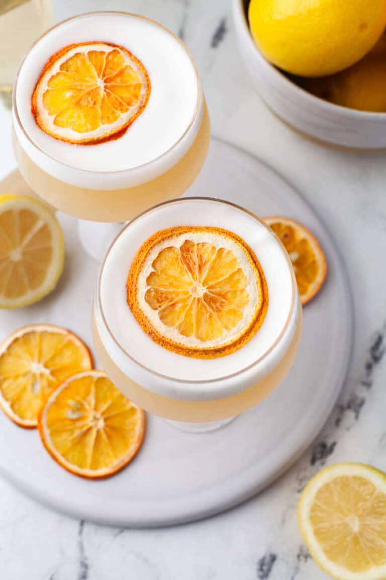Two coupe glasses filled with a frothy rum sour cocktail, each garnished with a dried orange slice, sit on a white plate. Slices of dried and fresh lemon surround the glasses, with a bowl of fresh lemons in the background.
