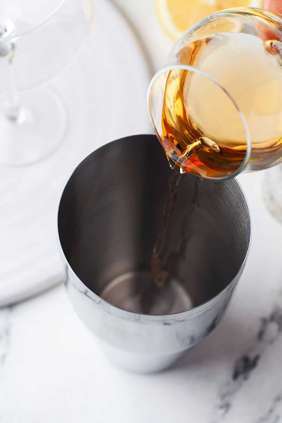 A close-up of a drink being poured from a glass into a stainless steel cocktail shaker, with part of a lemon and an empty glass visible in the background on a white surface.