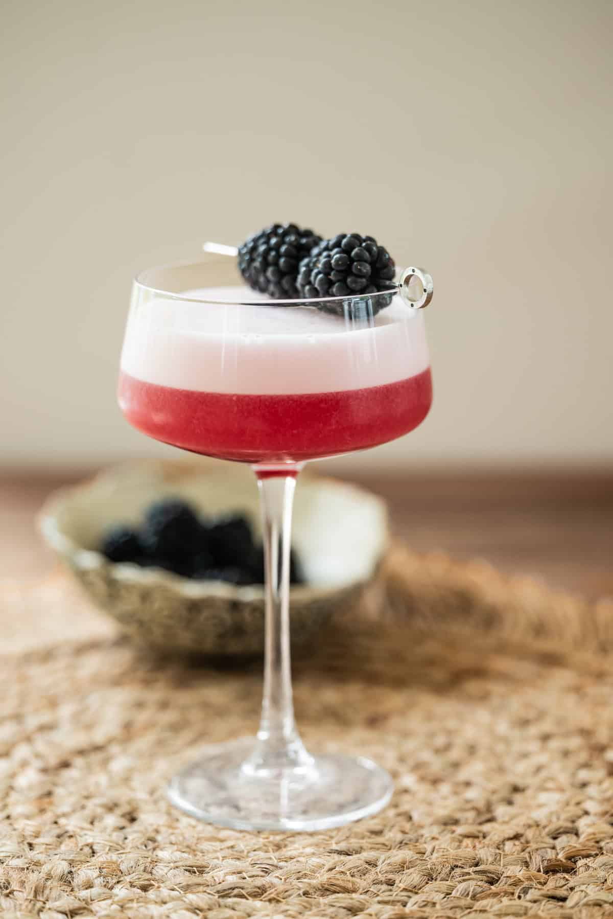A cocktail with a foamy pink layer, garnished with two blackberries on a pick, served in a coupe glass on a woven placemat. A bowl of blackberries is in the background.